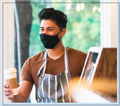 Male Barista With a Mask on Holding a Coffee Cup in Hand